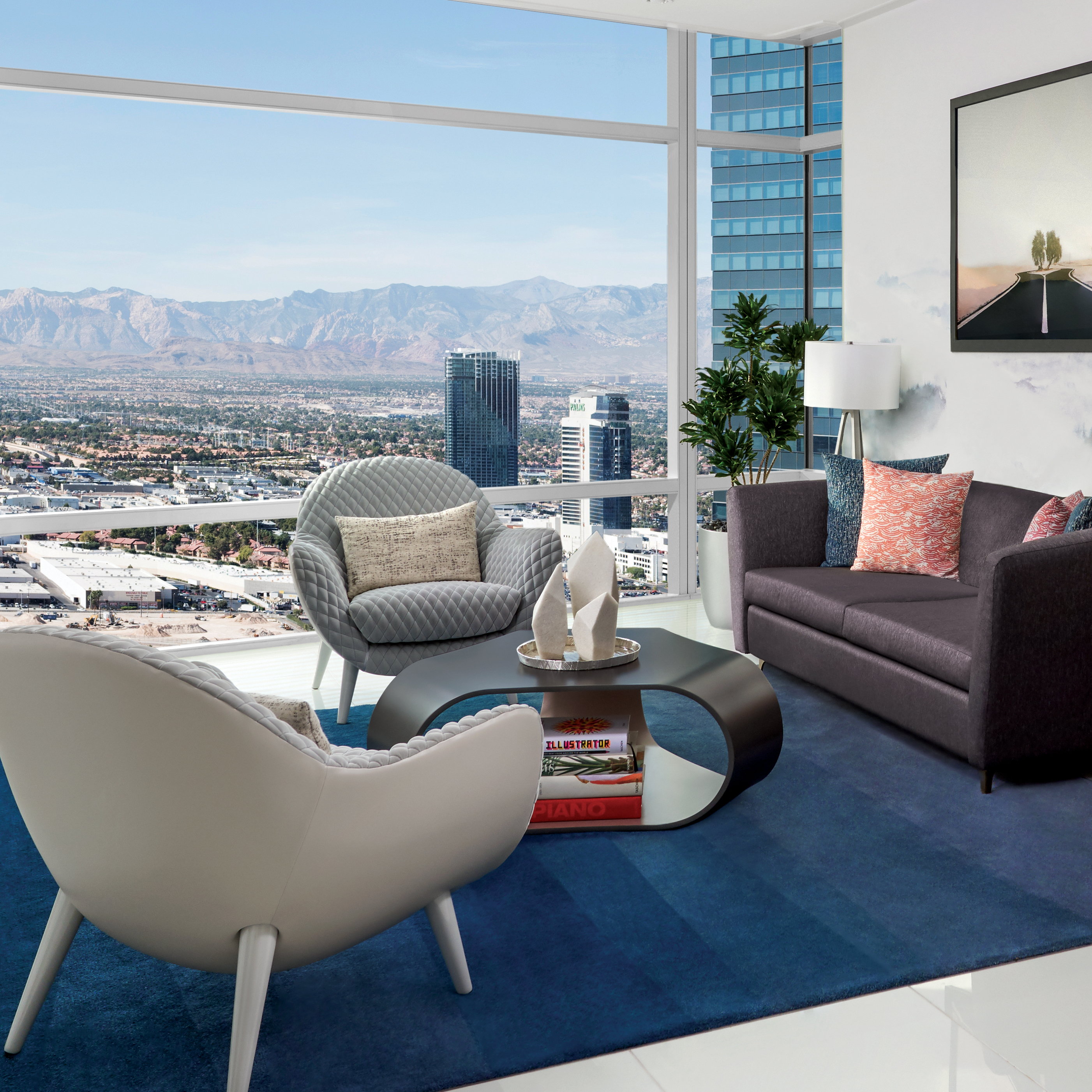 A modern hotel living room with a couch, armchair, and coffee table arranged in a seating area facing large floor-to-ceiling windows. The windows offer a panoramic view of a city skyline and mountains in the distance.
