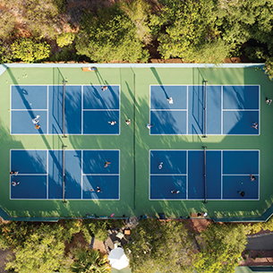  An aerial view of a group of people playing tennis on a bright green tennis court. The court is surrounded by tall trees with full green leaves. 