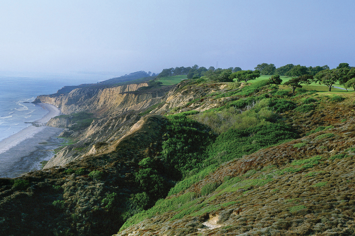 A golf course on a rocky cliff overlooking the ocean. The cliff is green with shrubs, grass and many trees.