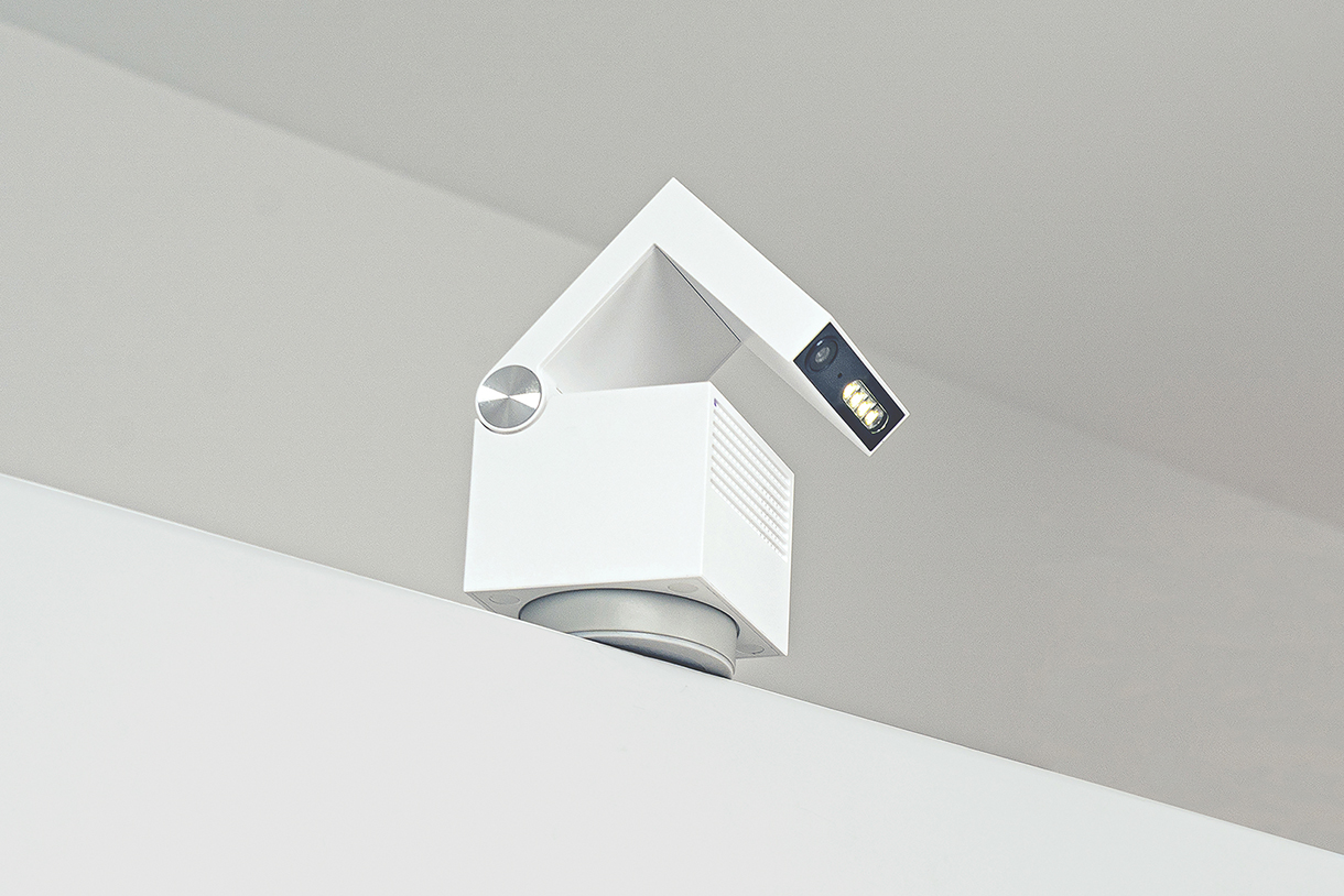 A small, white, cube-shaped projector with a tilting arm and a lens. It is mounted on a shelf.