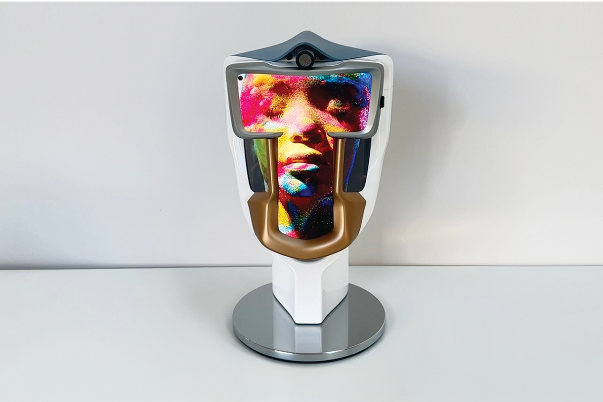 A futuristic, white head-shaped device with a gold-colored faceplate. A holographic image of a woman's face appears within the device.