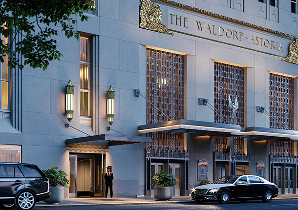 The exterior of the historical Waldorf Astoria hotel in New York City, a large Art Deco building with a doorman standing at the front entrance and two cars parked in front of it.