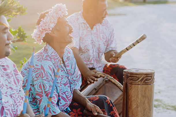 Three native Polynesian musicians playing traditional wooden drums. They are wearing colorful button-up aloha shirts with tribal and floral motifs. 