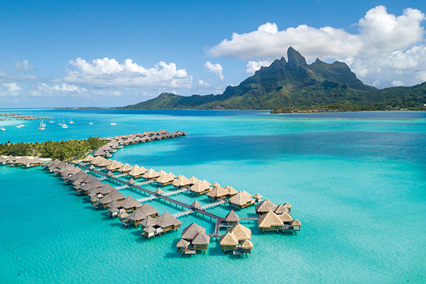 An aerial view of a luxury resort with overwater bungalows in the center of the South Pacific Ocean. The resort is located on a small island with a mountainous background. There is a motu, a small islet made of white sand, visible in the foreground.