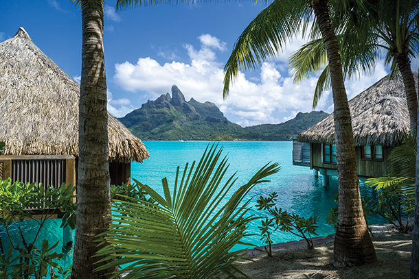 Two private thatched-roof bungalows are sitting above a large, tranquil, turquoise lagoon. In the foreground there are palm trees and in the background there are lush green mountains and blue sky.