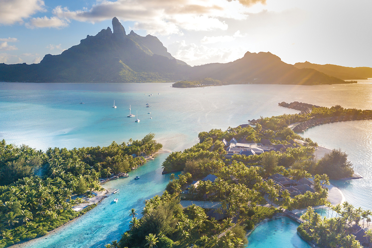 An aerial view of a tropical island with turquoise water, surrounded by a coral reef. In the background, lush green mountains rise up from the center of the island. There are several boats in the water and a resort on the island.