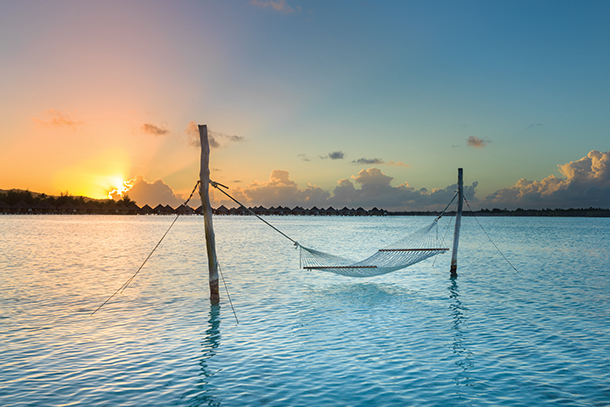 A hammock strung between two standing branches over a calm ocean at sunset. The sky is ablaze with orange, pink, and purple hues.