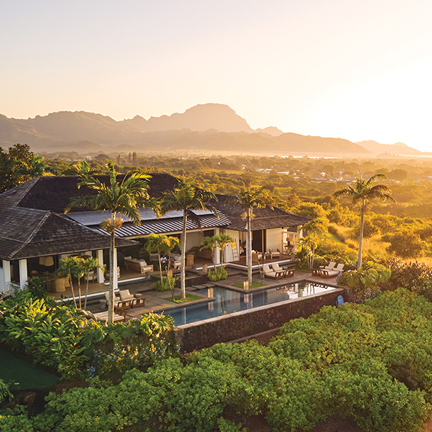 Aerial view of a modern luxury home with a pool, set against a backdrop of rolling hills and a distant mountain range at sunrise in Kauai.