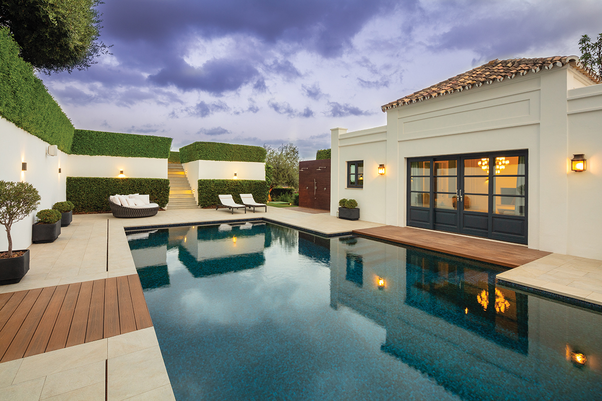 A modern villa with a swimming pool at dusk. The villa has white walls and a large retractable patio wall, with a wooden deck surrounding the pool. There are lounge chairs and potted plants around the pool.