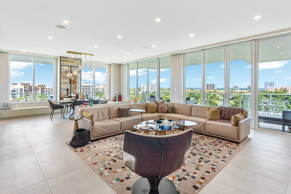 Spacious and modern living room with floor-to-ceiling windows offering panoramic ocean views. The room features a neutral color palette with beige sofas, a patterned rug, and a dining table with chairs.