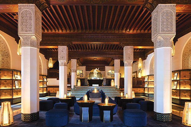 The lobby of the Mamounia Hotel in Marrakech, Morocco. The lobby features a high ceiling with ornate wood beams, arched columns, and decorative wall patterns. There are comfortable blue armchairs and small tables arranged around a central area with a fireplace.