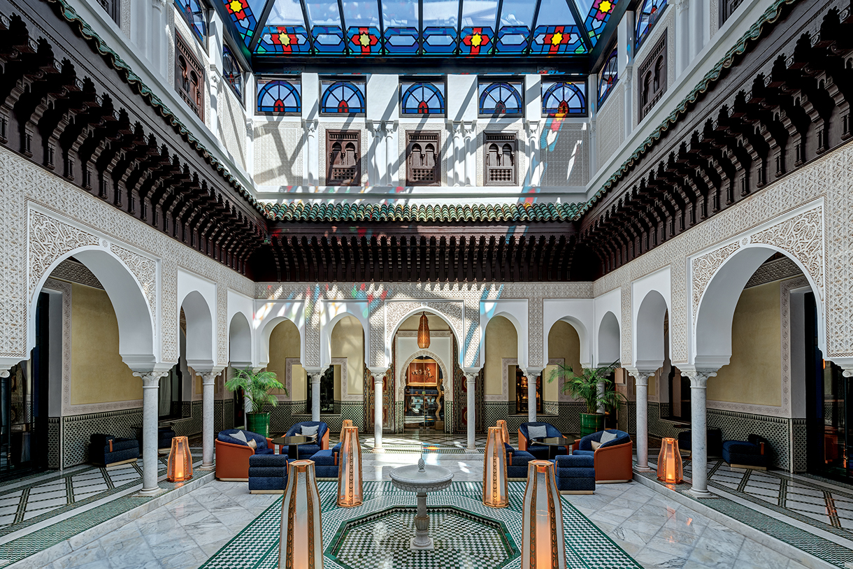 Interior shot of La Mamounia Hotel in Marrakech, Morocco. The image shows a large, open-air tea room and courtyard with a central fountain surrounded by arched columns and white walls. There are stained glass windows above and comfortable seating areas with blue armchairs and patterned rugs.