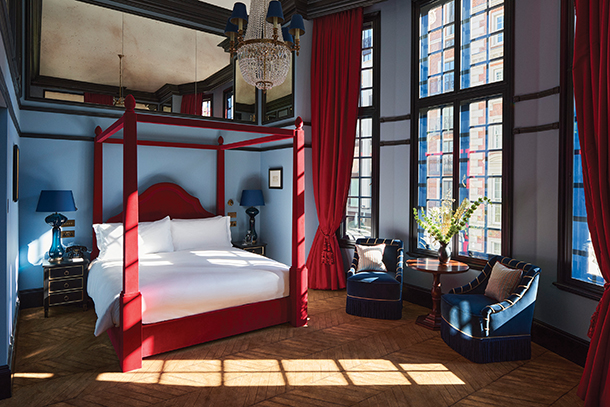 A luxurious hotel room with a four-poster bed, velvet curtains, and a chandelier. The room has large windows with city views and a sitting area with armchairs and a coffee table.