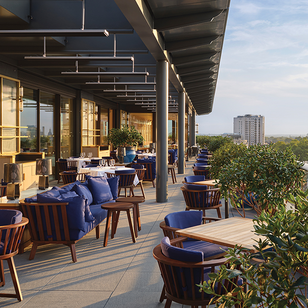 A rooftop terrace with outdoor seating, featuring blue armchairs, wooden tables, and potted plants. The terrace overlooks a cityscape with buildings and trees.