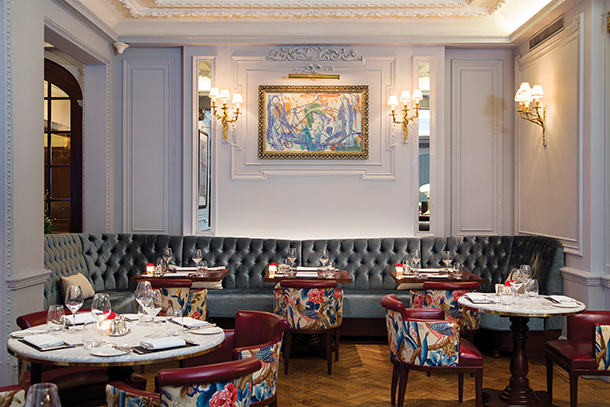 A stylish, yet classy restaurant with a large blue gray upholstered bench and round marble and wood tables. The chairs have colorful floral patterns and there is an abstract wall painting hanging.