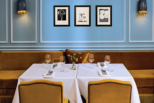 A beautiful dining room with blue walls and a white tablecloth on a table. There are yellow upholstered chairs, wine glasses,and a small vase of flowers. Three framed pictures hang on the wall.