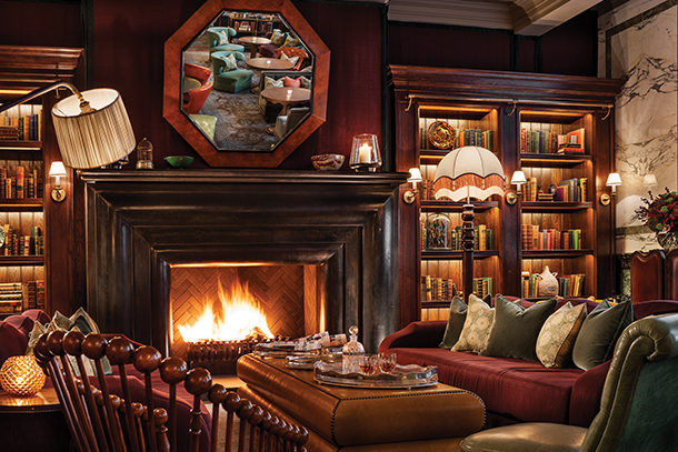 A cozy, inviting bar lounge with a fireplace, bookshelves, and comfortable seating. The room is decorated in rich, warm tones with dark wood accents and colorful pillows.