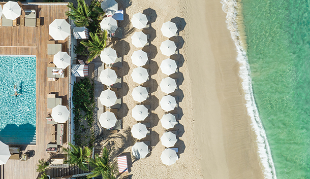 An aerial view of the Cheval Blanc St-Barth beach front resort property with a swimming pool, large wooden deck, sandy white beach with white umbrellas and vibrant turquoise ocean. 