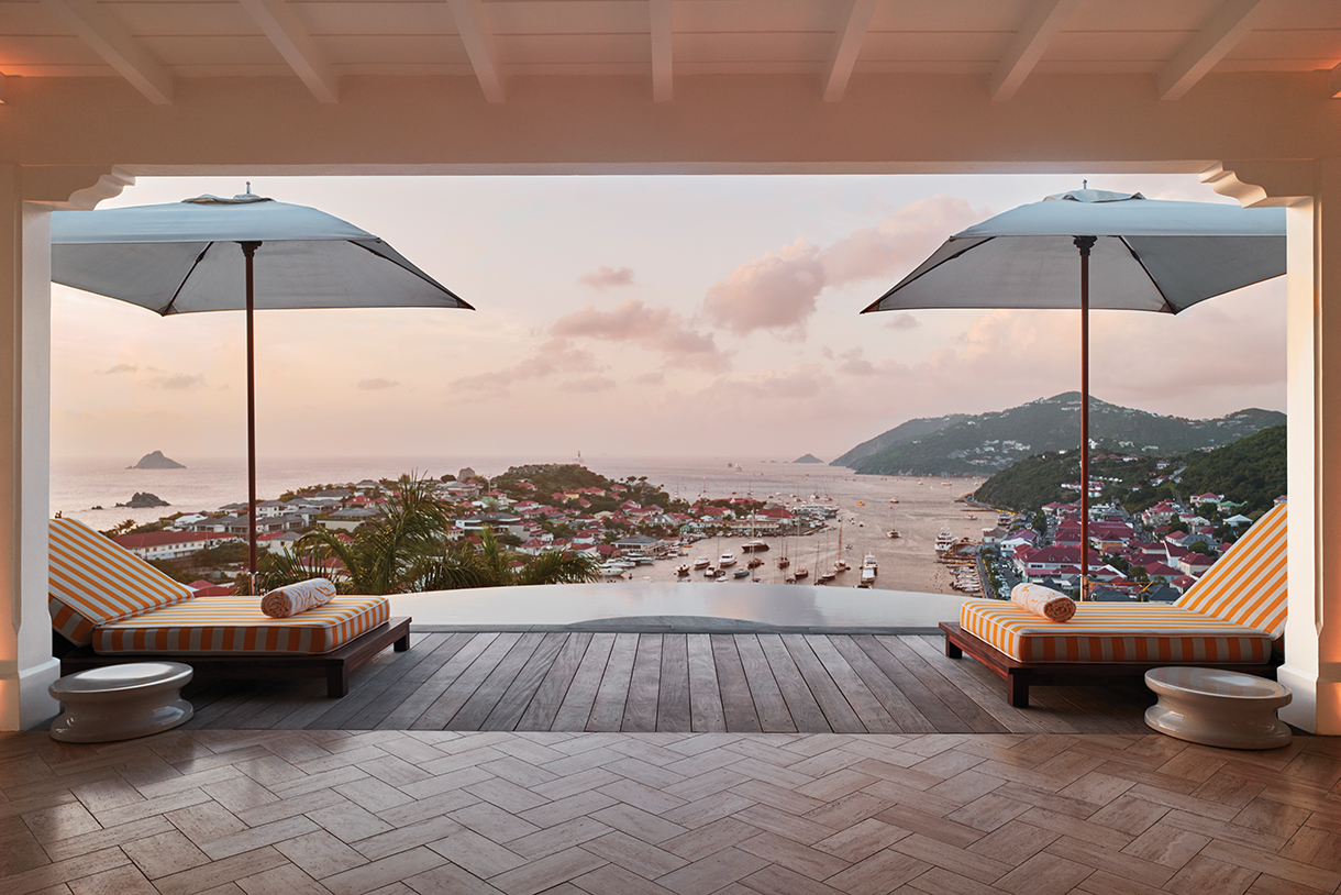 A luxurious hotel patio deck with lounge chairs and umbrellas located on a hilltop, overlooking the ocean with sailboats visible in the distance. 
