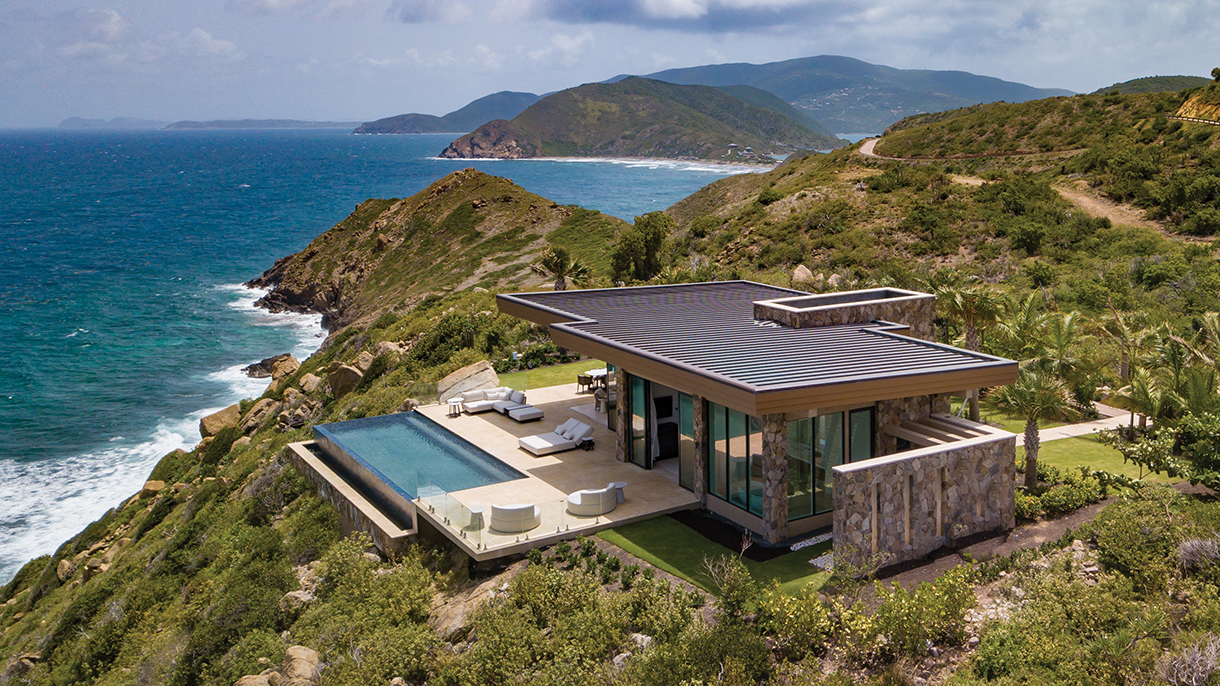 A luxurious, modern resort property with a swimming pool built on a cliff overlooking the Caribbean Ocean. The house has large windows that provide panoramic views of the water.