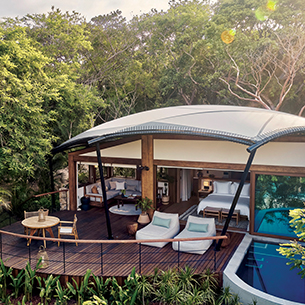 An open-air living room of a large resort property built on a platform overlooking lush jungle. The large tented room has floor-to-ceiling windows and a deck with a private plunge pool, table and lounge chairs.