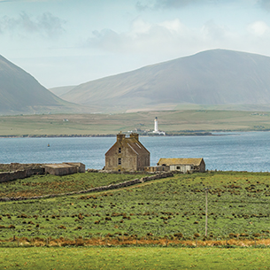 An old house located on the coast surrounded by grass with ocean and mountains in the background.