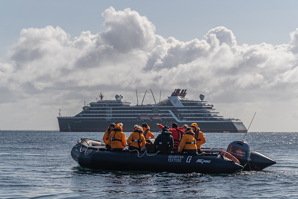 Passengers wearing orange life jackets and safety gear aboard a black rubber Zodiac  boats in the Celtic Sea. The Seabourn Venture is sailing in the background.