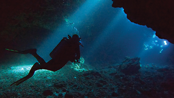  A silhouette of a scuba diver exploring a dark underwater cavern. The diver is illuminated by a beam of sunlight entering from above.