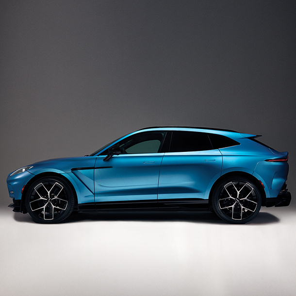 A side view of a blue Aston Martin DBX SUV parked in a studio or showroom. The SUV has black wheels and a luxurious, modern, and sleek design.