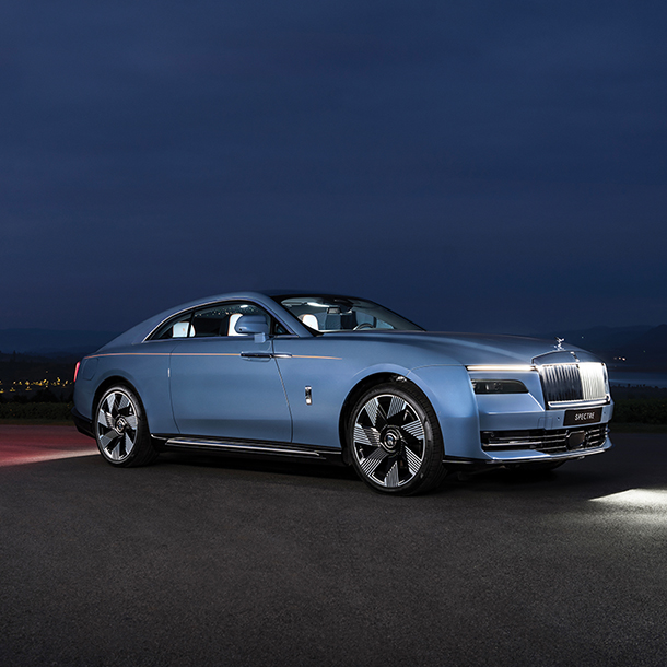 A front side view of a blue Rolls-Royce Spectre parked on the side of a road at night.)