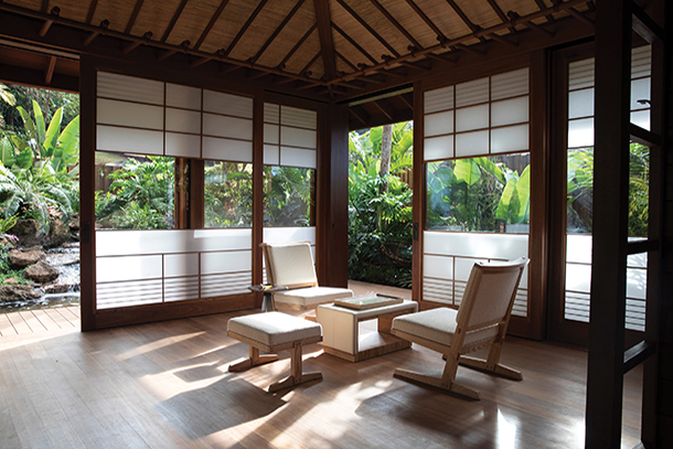 A serene and minimalist room with sliding shoji screens, wooden floors, and comfortable seating, overlooking a lush tropical garden.