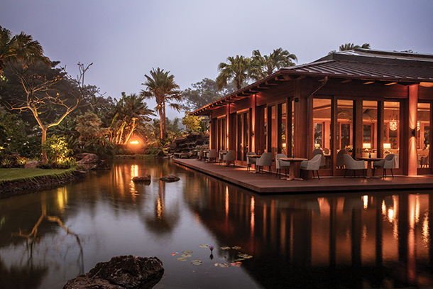 A serene restaurant building with a wooden deck overlooking a tranquil pond, illuminated by warm lights at dusk.