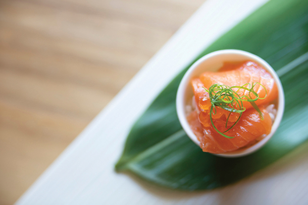 A close-up of a bowl of fresh salmon sashimi, topped with salmon eggs and green onions, served on a bed of rice and a large green leaf.