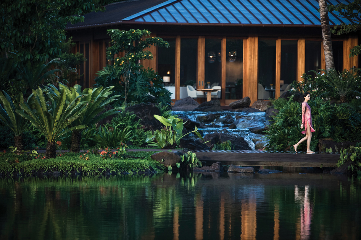 A woman in a pink dress walks along a wooden bridge over a serene pond at Sensei Lanai, a luxury wellness resort in Hawaii. Lush greenery surrounds the pond, and a waterfall cascades into it.