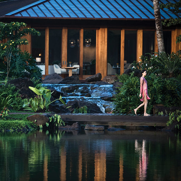 A woman in a pink dress walks along a wooden bridge over a serene pond at Sensei Lanai, a luxury wellness resort in Hawaii. Lush greenery surrounds the pond, and a waterfall cascades into it.