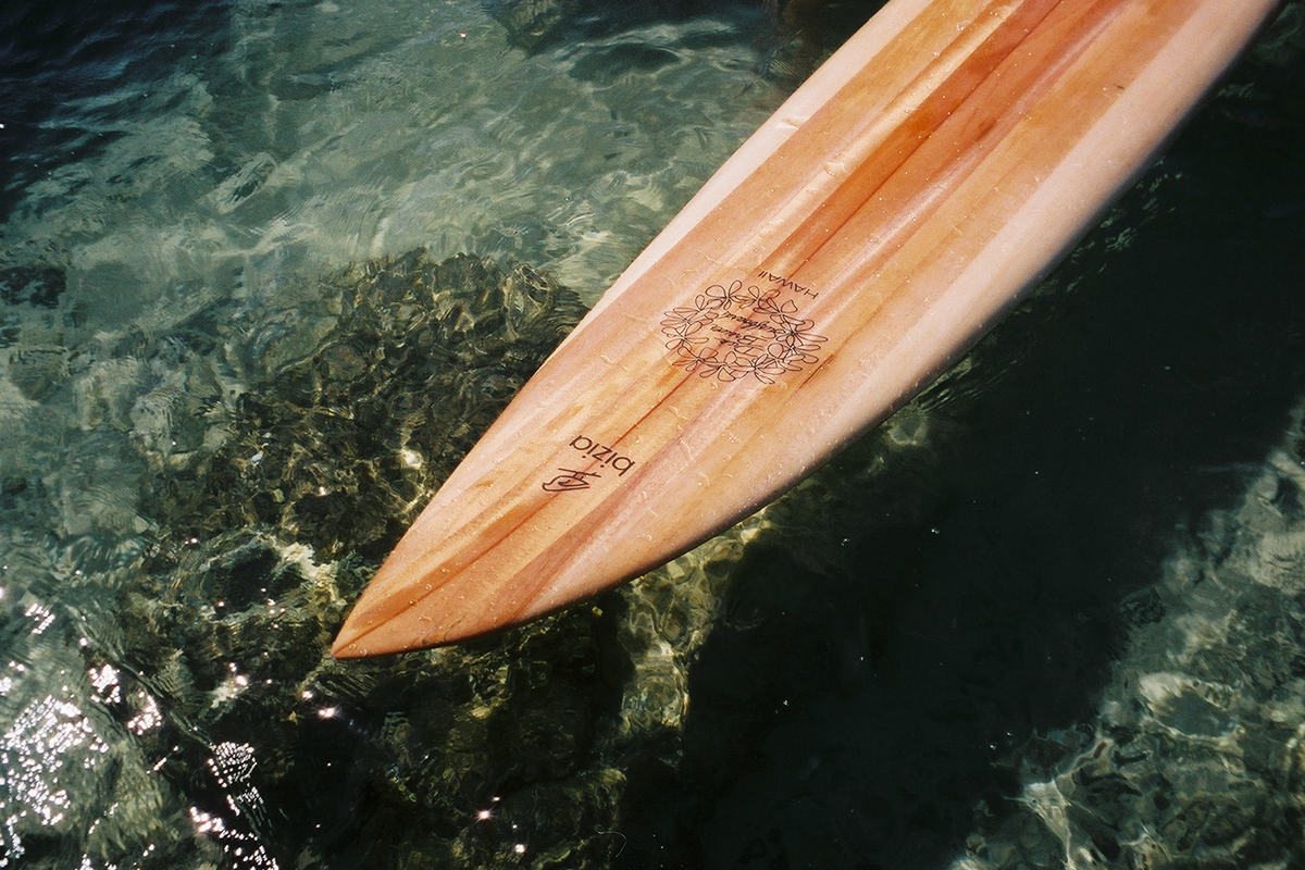 Strong, but lightweight wooden longboard on water. Longboard has a bird logo and plumeria lei on it and is handmade of hollowed-out wood, from Hawaiian albizia tree. 