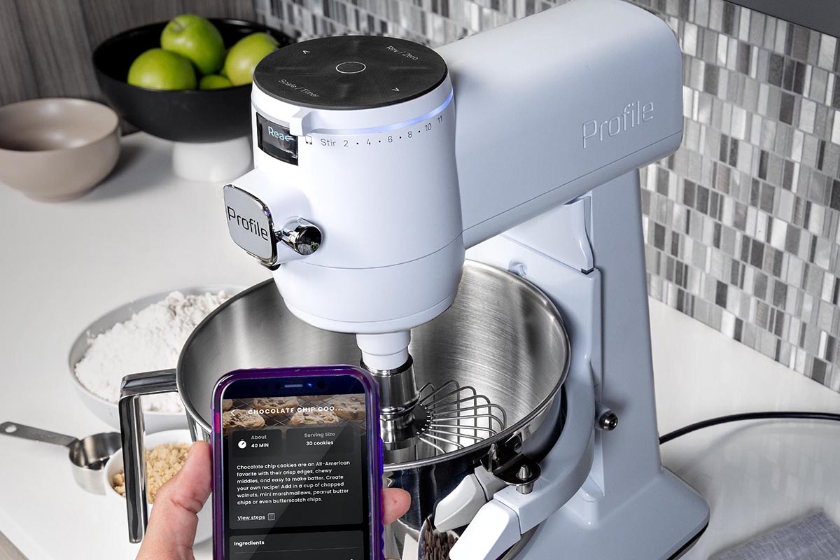 A person holding a cell phone with a recipe in front of a GE Profile white stand mixer. The mixer is on a white countertop with gray and white blacksplash.