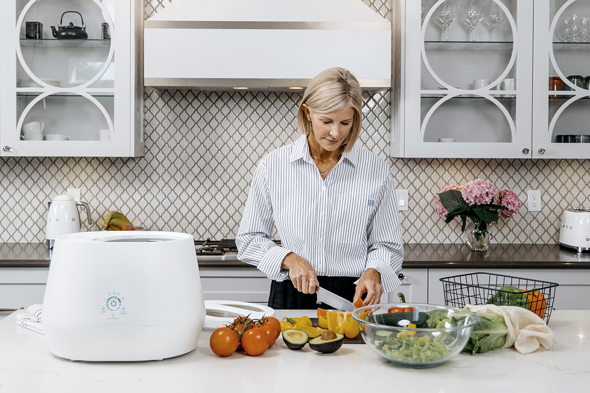 Woman in a kitchen cutting vegetables on a white kitchen island. The countertop has a white food-waste composter sitting next to tomatoes, peppers and avocados.