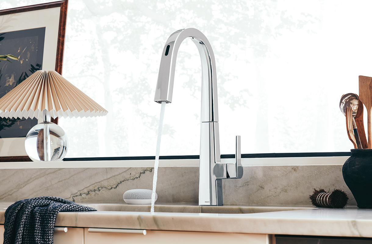 A kitchen sink with water running from a chrome faucet. The sink has a natural stone counterop and backsplash, a dark towel hanging from it, and a window above it.