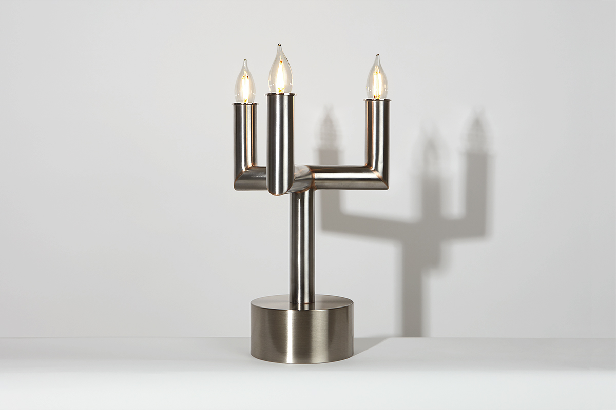 The Candle Table Lamp from Blue Green Works is a modern take on the candelabra, featuring a minimalist, triple-armed design crafted of stainless steel on a white background.