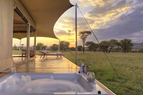 Mobile Bushtops Safari Camps set up in the middle of a grassy field. The tent has large side openings, with a bed, table and chairs with view of Tanzania's wildlife. 