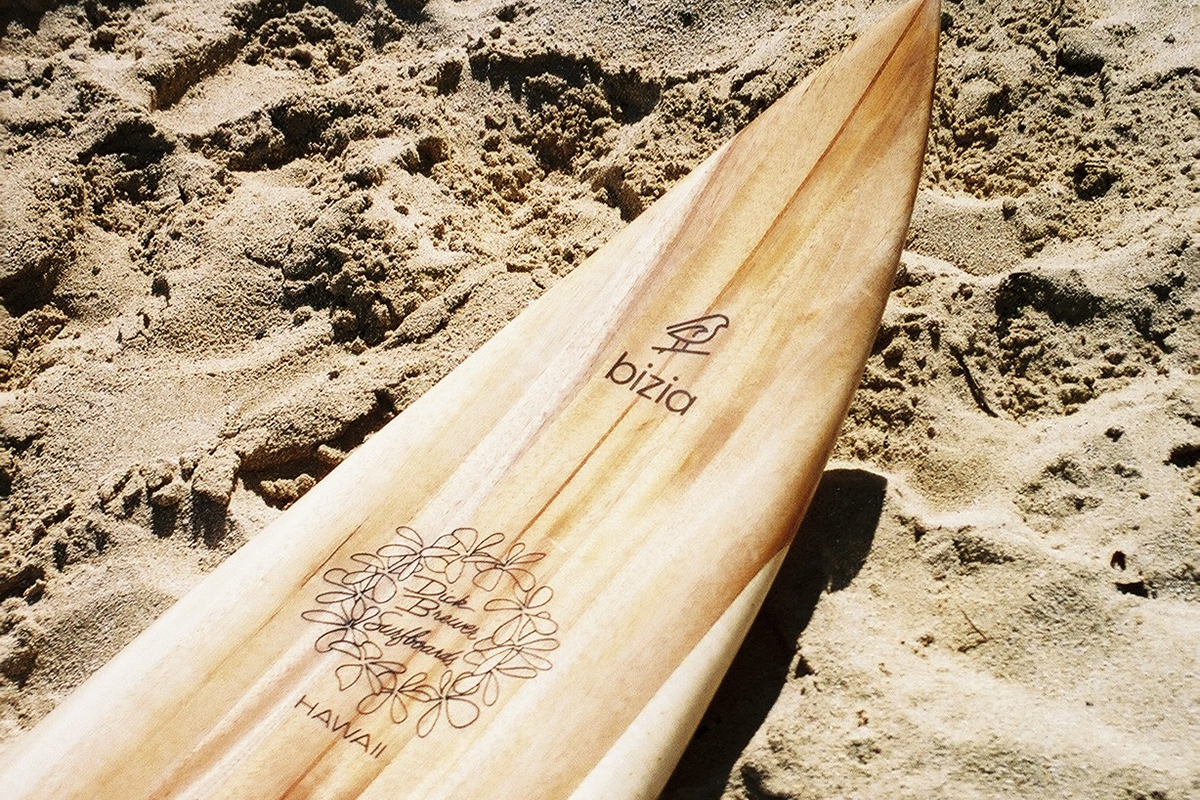 Strong, but lightweight wooden longboard on sand. Longboard has a bird logo and plumeria lei on it and is handmade of hollowed-out wood, from Hawaiian albizia tree. 