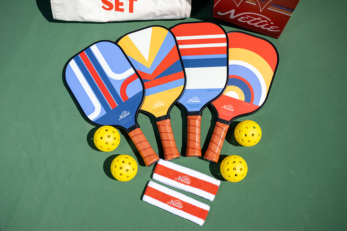 Nostalgic Pickelball set including 4 colorful, retro-patterned paddles, 4 red-striped headbands and 4 yellow pickleballs placed on a green background. 