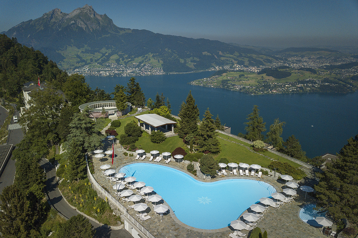 Aerial view of a large swimming pool with lounge chairs at a Bürgenstock Hotel & Spa surrounded by lush greenery overlooking a beautiful lake on a mountain top.