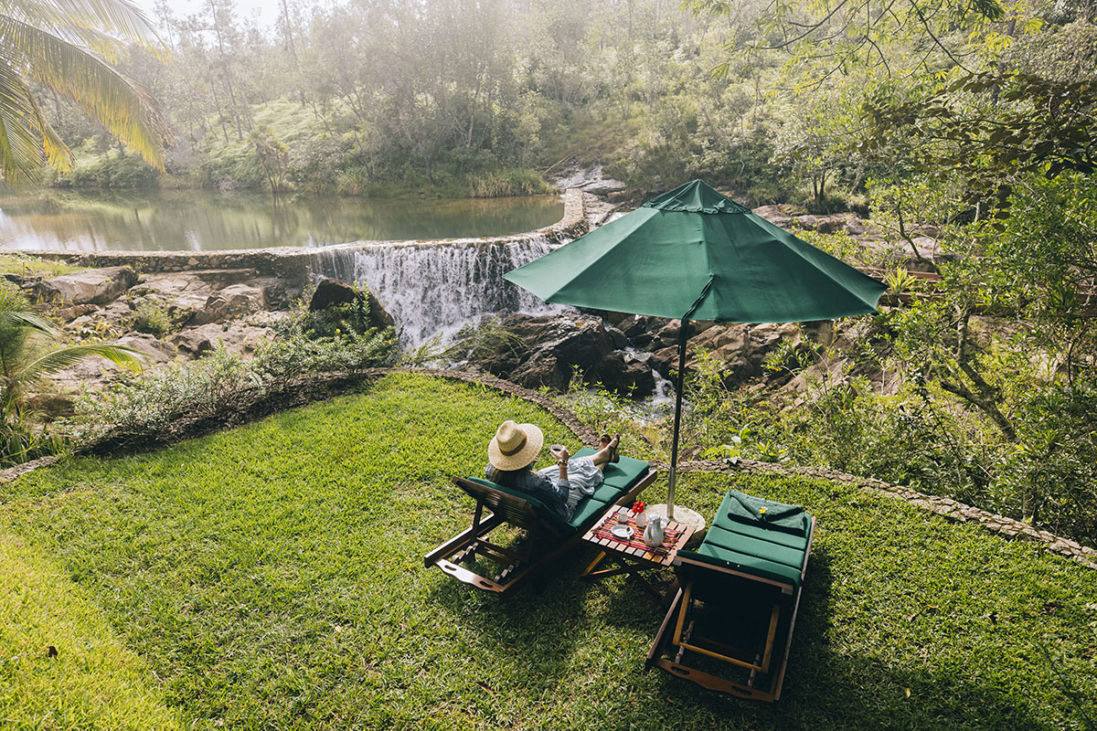 A woman sitting in a lounge chair under a green umbrella in front of a large and powerful waterfall at the Blancaneaux Lodge part of the Family Coppola Hideaways. 