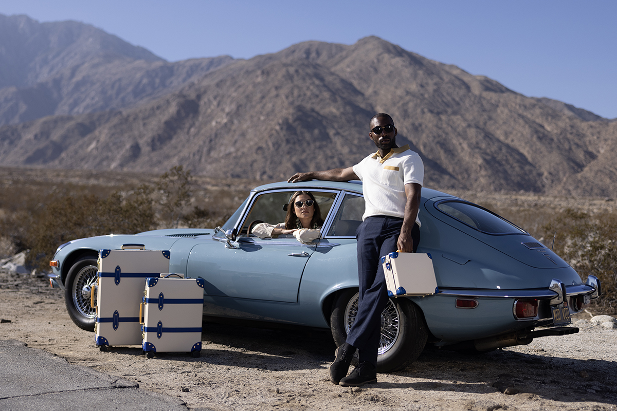 A man standing next to a blue vintage car in the desert with a woman sitting inside. He is carrying Globe-Trotter luggage and there are additional luggage on the ground. 