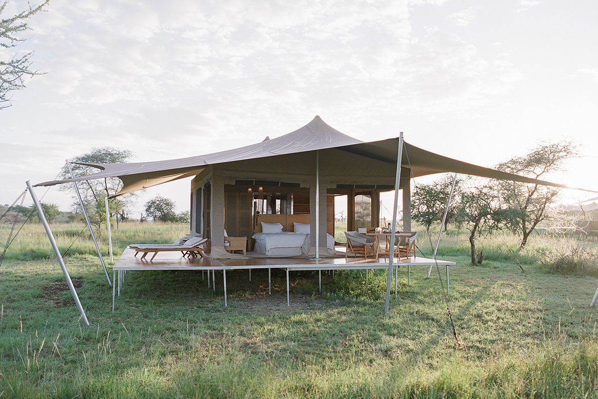 Mobile Bushtops Safari Camps set up in the middle of a grassy field. The tent has large side openings, with a bed, table and chairs with view of Tanzania's wildlife. 