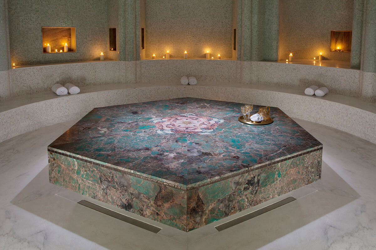 A luxurious spa room with a stunning, large marble hammam or Turkish bath in the center. The hammam is surrounded by marble benches and illuminated by warm, soft lighting. 