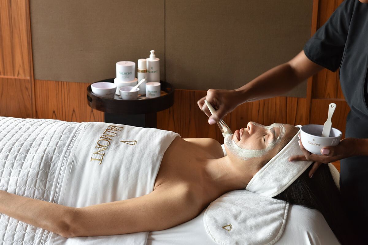 A person lying on a spa table with a white towel covering their body and a therapist is performing a facial treatment. The therapist is applying a white cream to the person's face. The background shows a luxurious spa room with neutral colors and dim lighting.