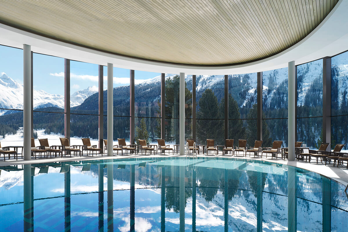 A luxurious indoor swimming pool area in a spa, with warm lighting and elegant decor. The pool is surrounded by lounge chairs. The view outside showacases a snowy mountain with large green trees.